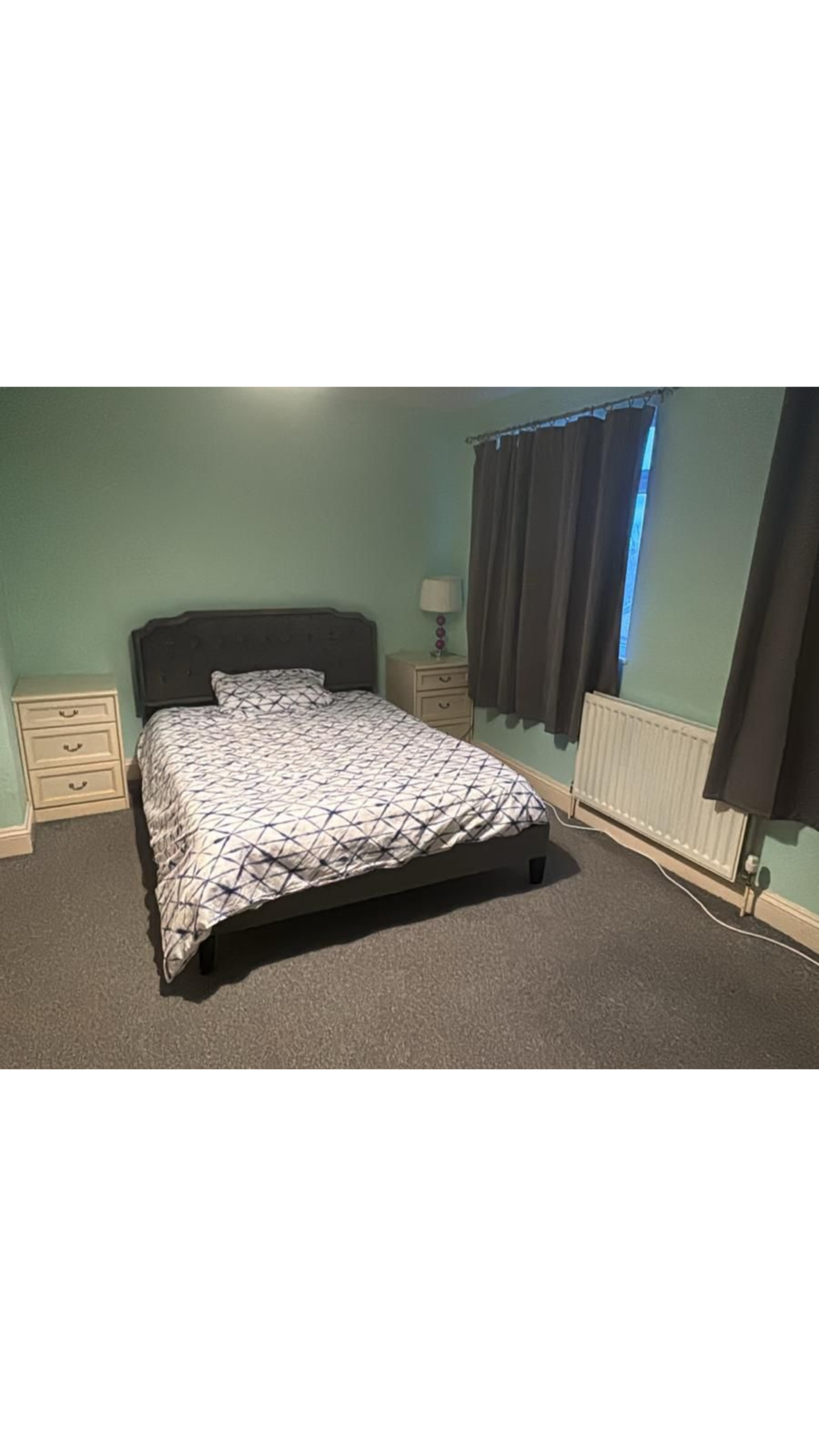 Bedroom with teal walls featuring a dark upholstered bed, white geometric duvet, and two beige nightstands with a lamp between them at the bedside.